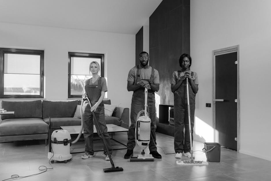 A black-and-white image of three professional cleaners in a modern living room with a light-colored tiled floor. The team includes two women and one man, all dressed in uniform workwear, standing with